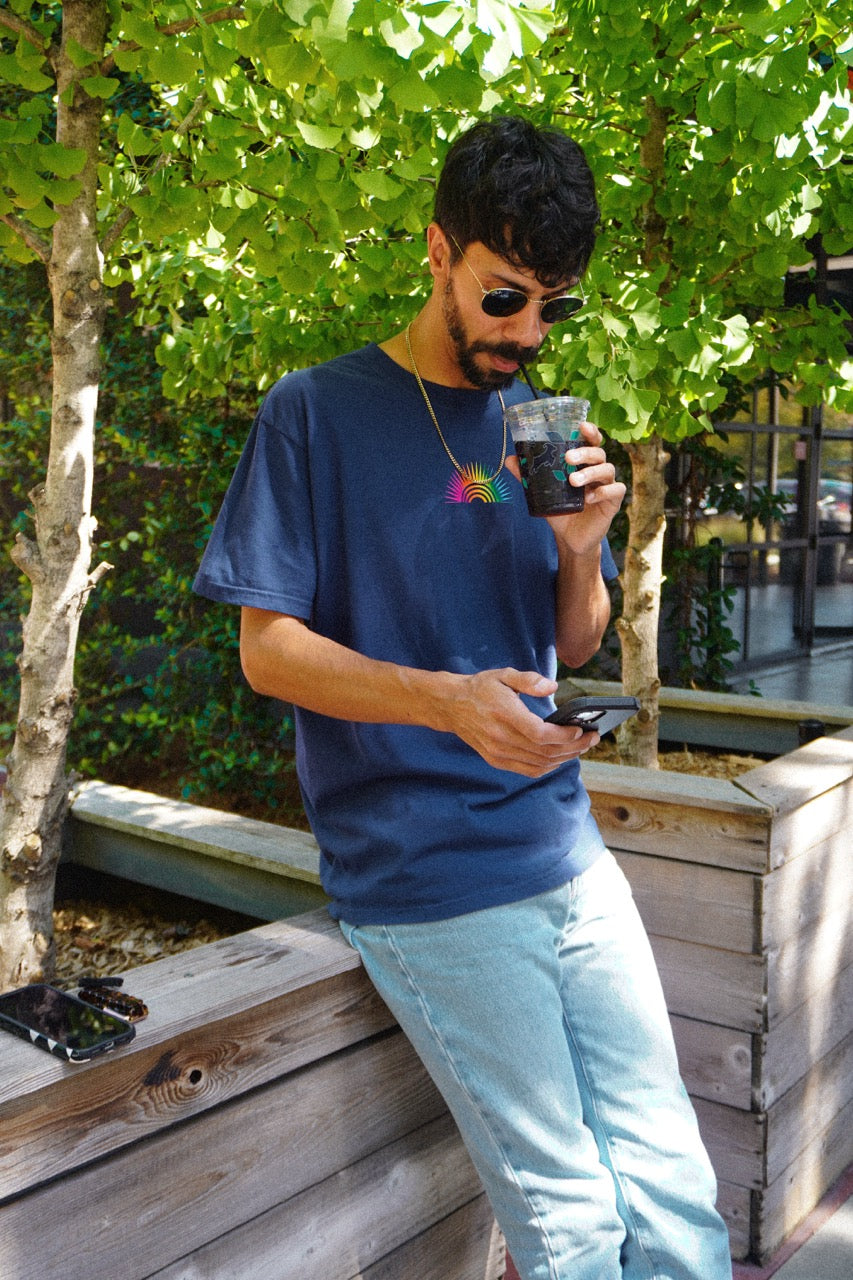 A man wearing a navy blue t-shirt with a colorful rainbow graphic on the chest, light blue jeans, and sunglasses, sitting on a wooden bench outdoors and holding a drink.