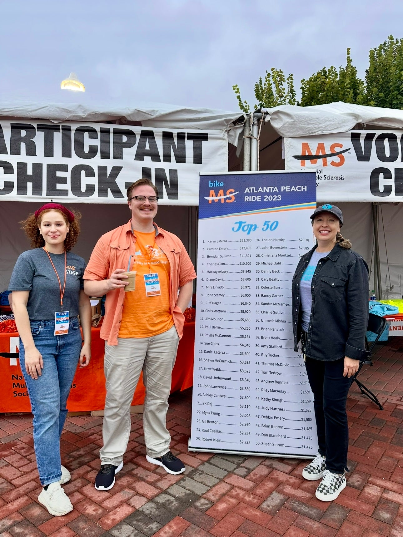 Three people standing in front of a 'Participant Check-In' sign at an event with a banner listing top participants.