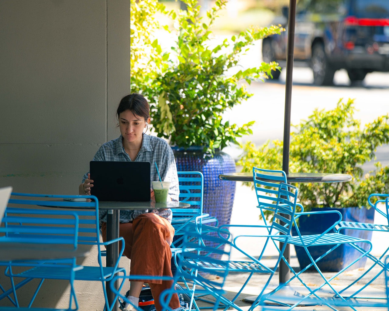 Person using a laptop at an outdoor cafe with blue chairs and greenery.
