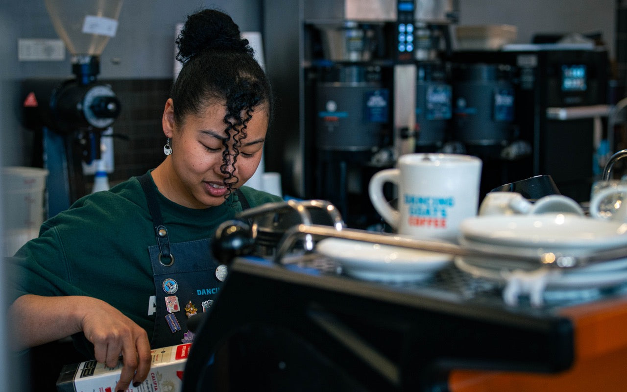 Person working behind a counter in a coffee shop with coffee equipment and cups.