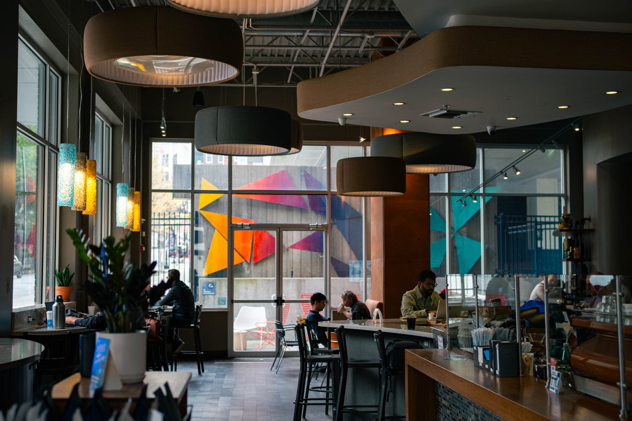 Modern cafe interior with large windows, colorful abstract art, and people sitting at tables.