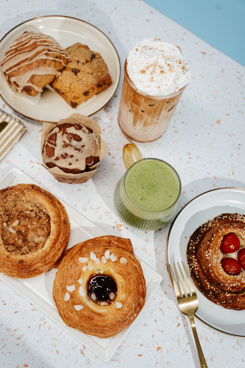An overhead styled food and beverage spread on a speckled white table, featuring pastries, a iced coffee with whipped cream, a green drink, and a pastry with red berries, arranged with plates and a gold fork.