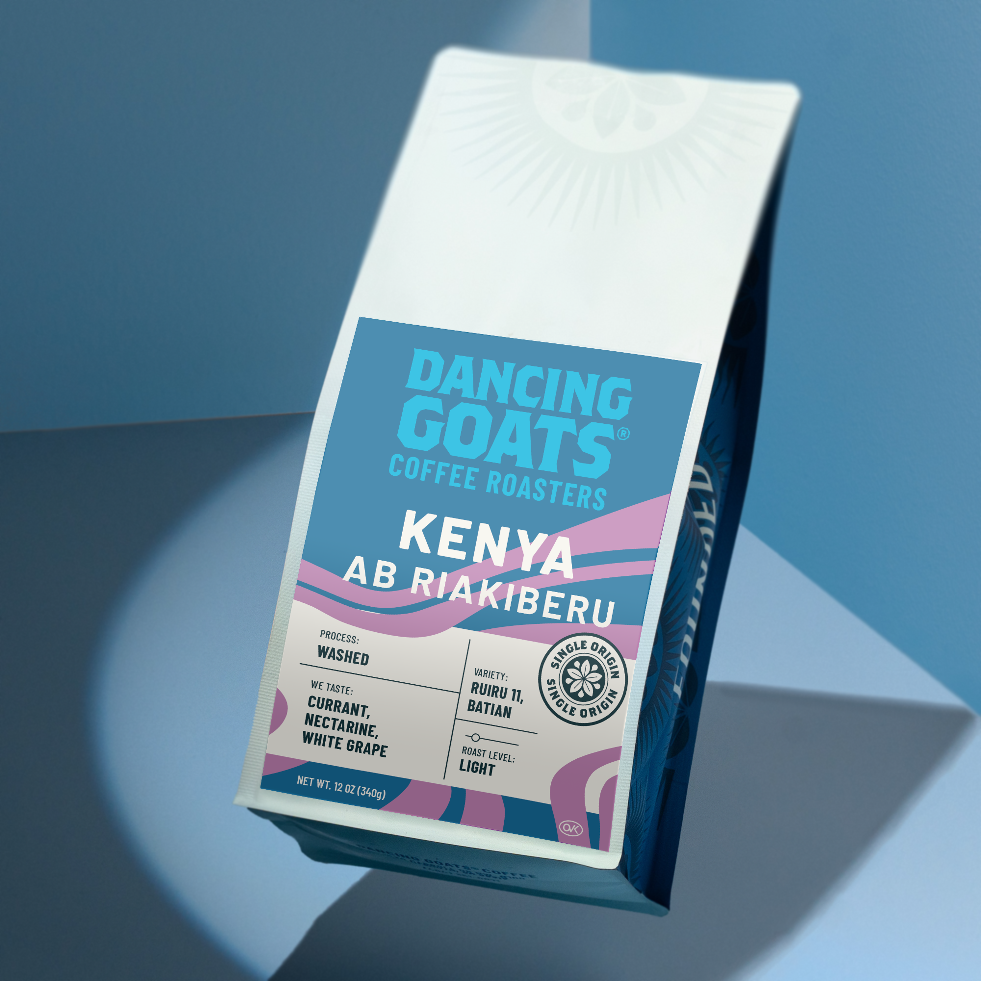 A bag of Dancing Goats Coffee Roasters Kenya Ab Riakiberu coffee, shown at an angle on a blue and gray geometric background. The packaging is white with a prominent blue label and pink accents.