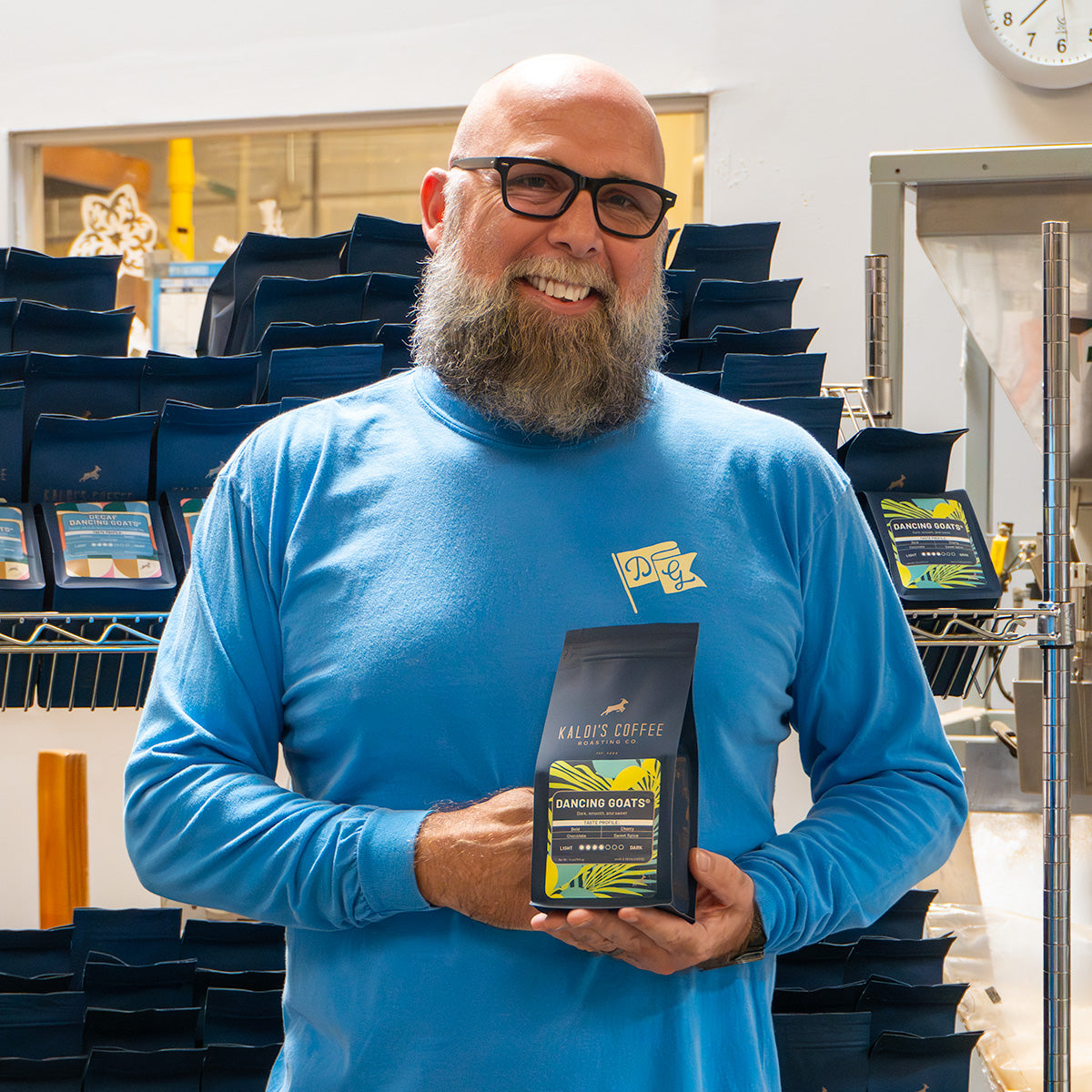 smiling man wearing blue long sleeve dancing goats shirt and holding bag of dancing goats blend coffee