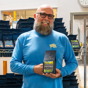 smiling man wearing blue long sleeve dancing goats shirt and holding bag of dancing goats blend coffee