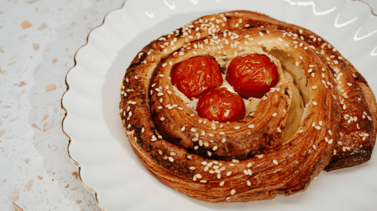 A spiral pastry with three halved cherry tomatoes in the center and white sesame seeds on top, served on a white scalloped plate.