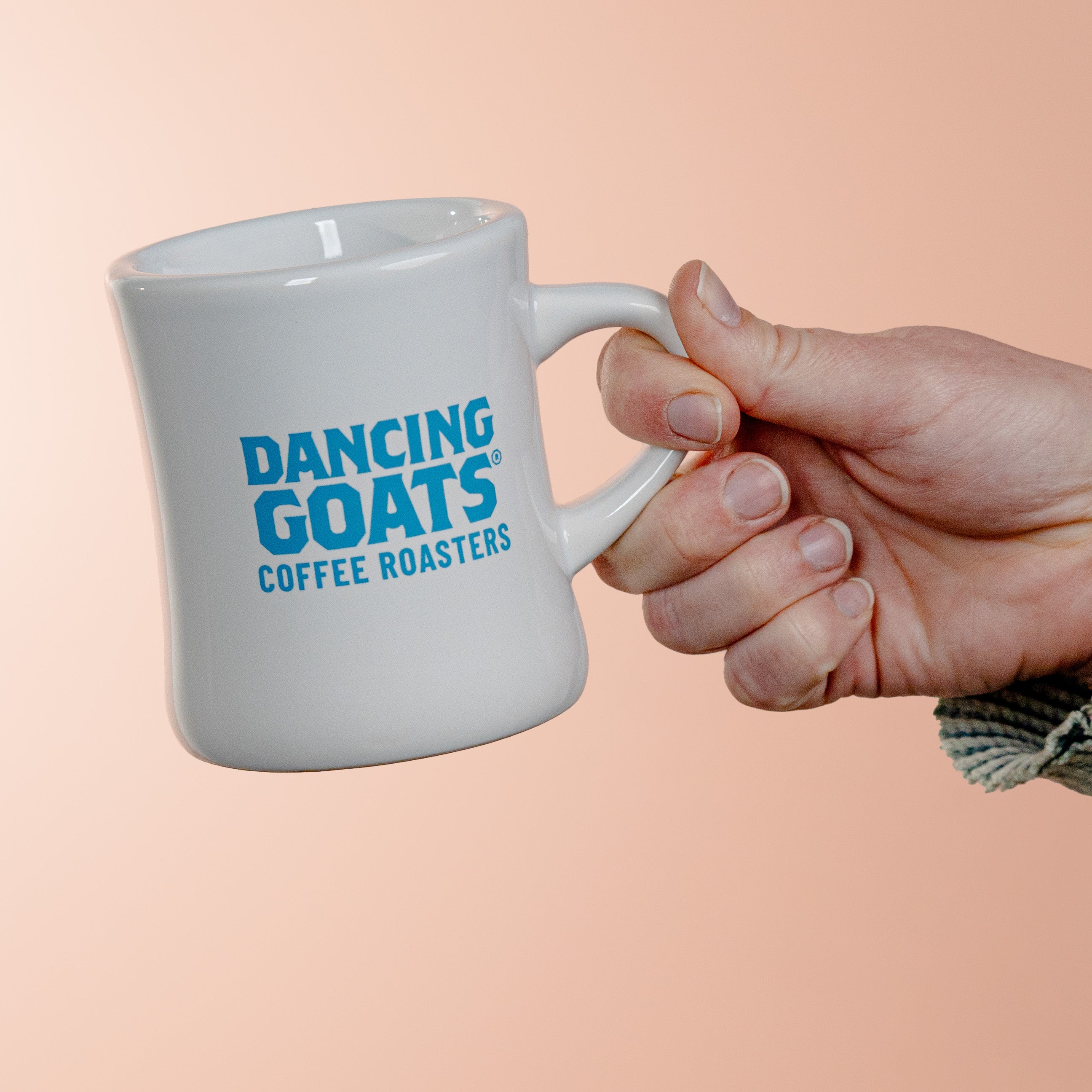 White mug with 'Dancing Goats Coffee Roasters' text held by a hand on a peach background
