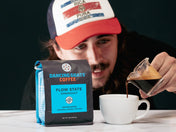 Person pouring coffee from a glass container into a white cup with a 'Dancing Goats Coffee' bag in the foreground.