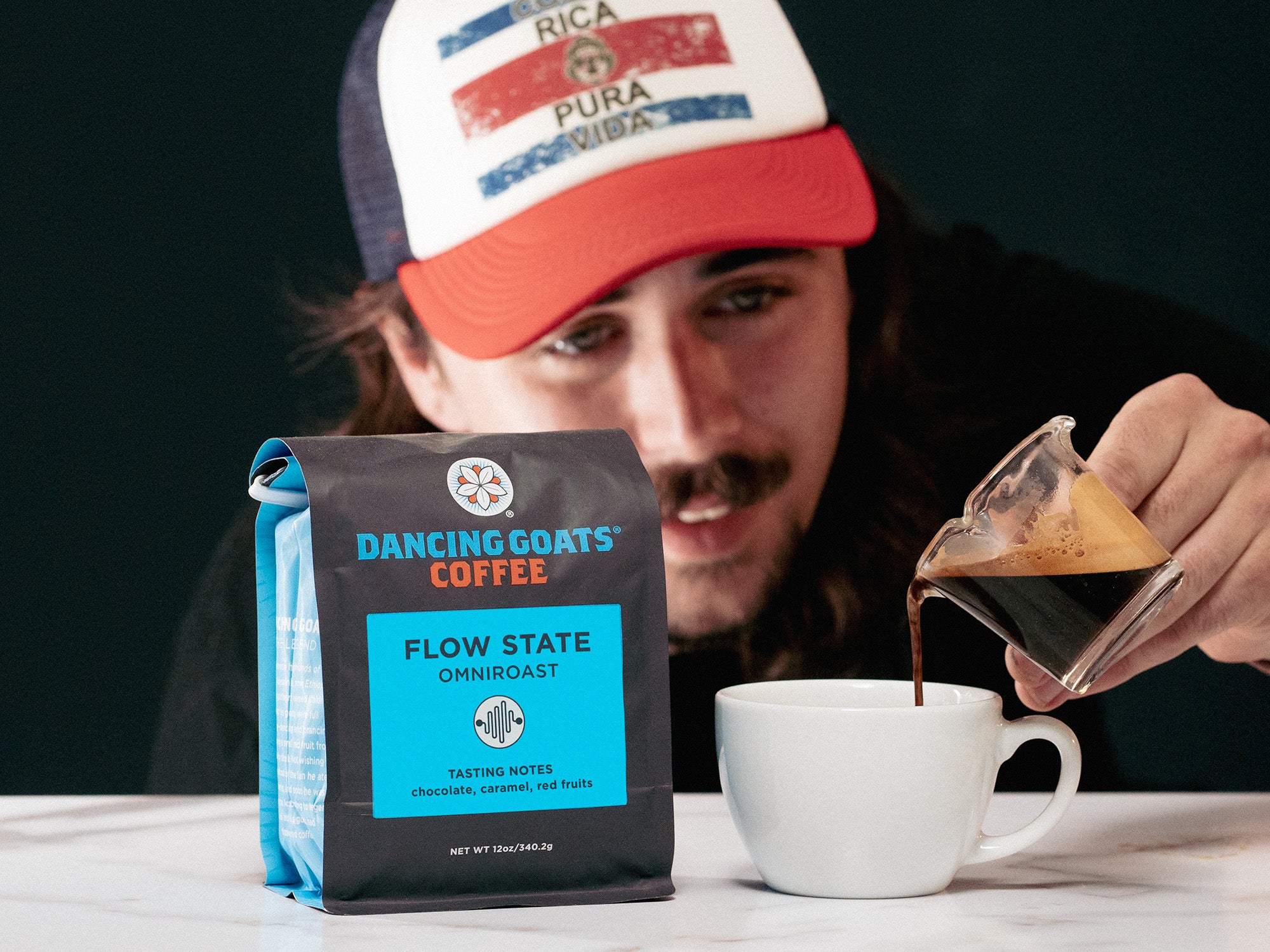 Person pouring coffee from a glass container into a white cup with a 'Dancing Goats Coffee' bag in the foreground.