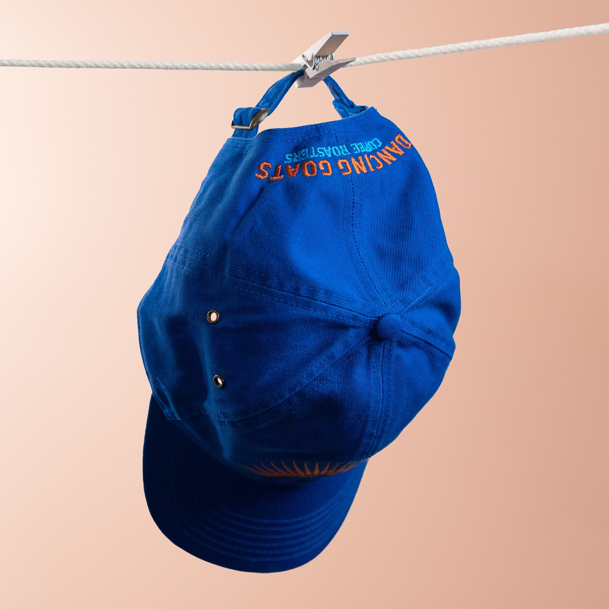 Blue cap with text: dancing goats coffee roasters, hanging on a clothesline against a beige background
