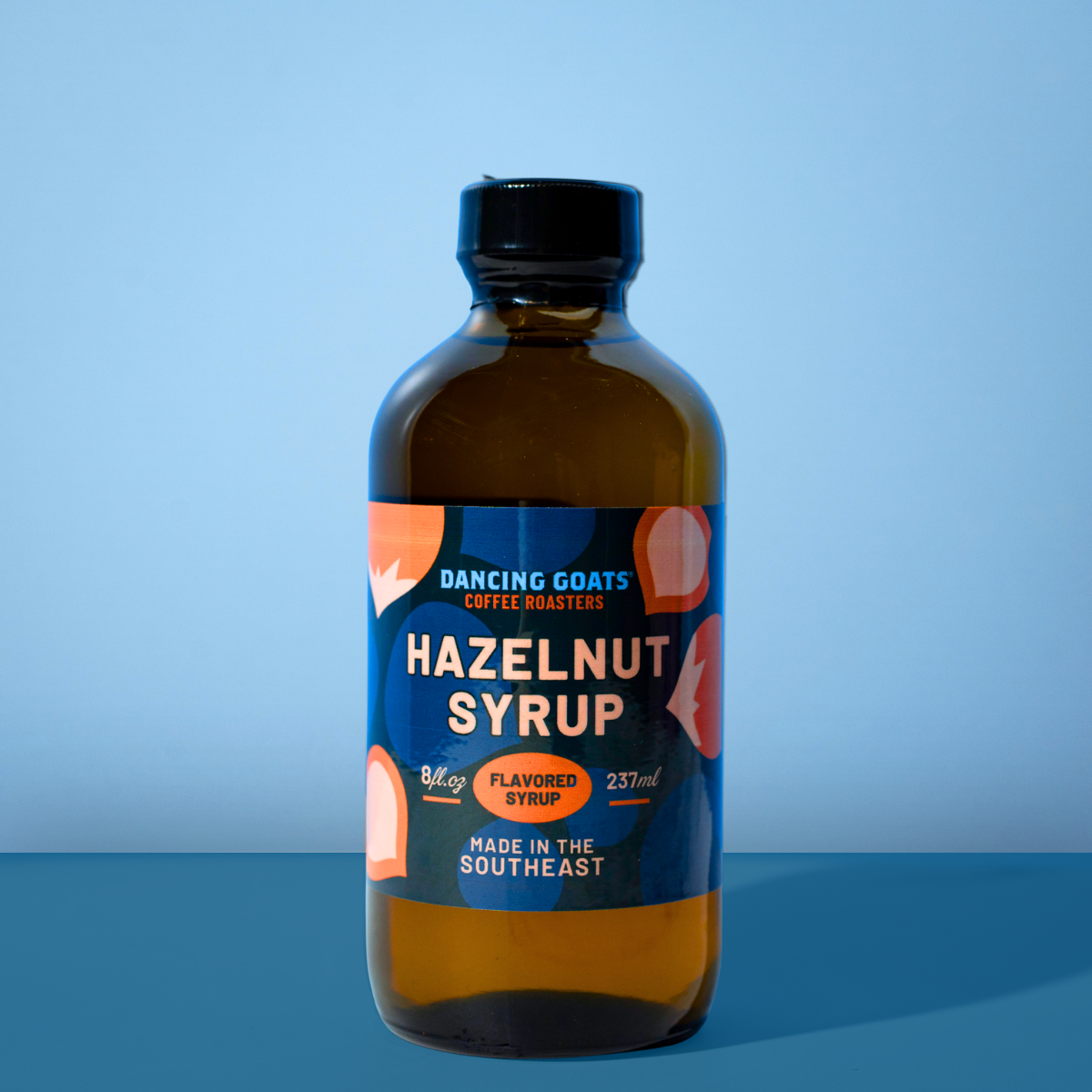 A bottle of Dancing Goats Coffee Roasters Hazelnut Syrup, 237ml, featuring an amber glass bottle with a black cap and a dark blue label with orange and white text, set against a solid blue background.