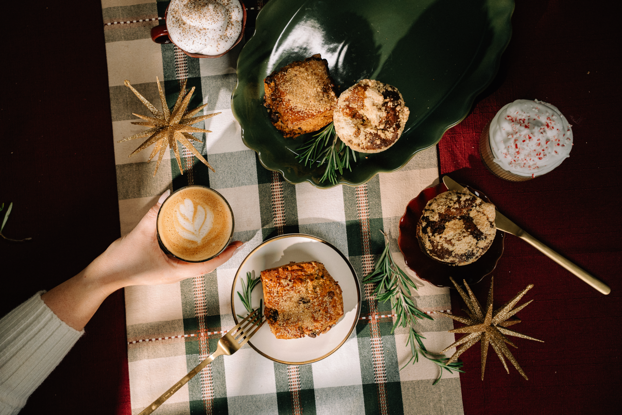 An overhead lifestyle shot of a cozy holiday table setting. A hand holds a cup of latte art coffee on the left, next to a plate of seasonal cake slices.