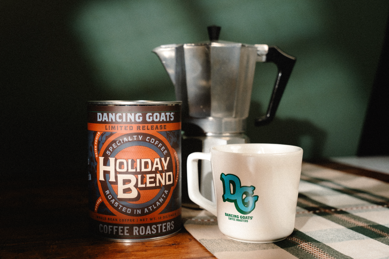 A can of Dancing Goats Holiday Blend roasted coffee sits on a wooden table next to a white ceramic mug with a teal logo, with a silver moka pot in the background against a dark green wall.