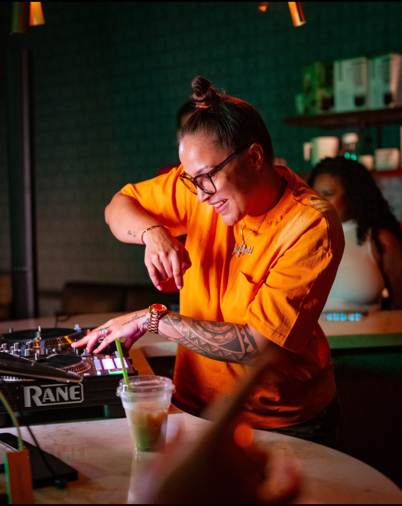Person in orange shirt using DJ equipment with a drink and straw in a bar setting