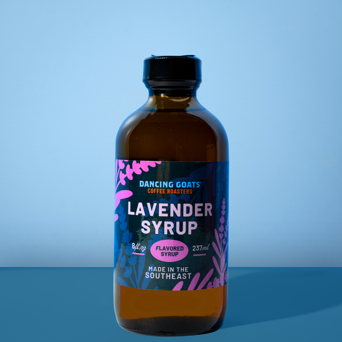 A bottle of Dancing Coats Lavender Syrup with a black cap and a dark blue label featuring pink floral illustrations, set against a solid blue background.