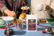 Person preparing coffee with a 'Holiday Blend' package on a table with snacks and a mug.