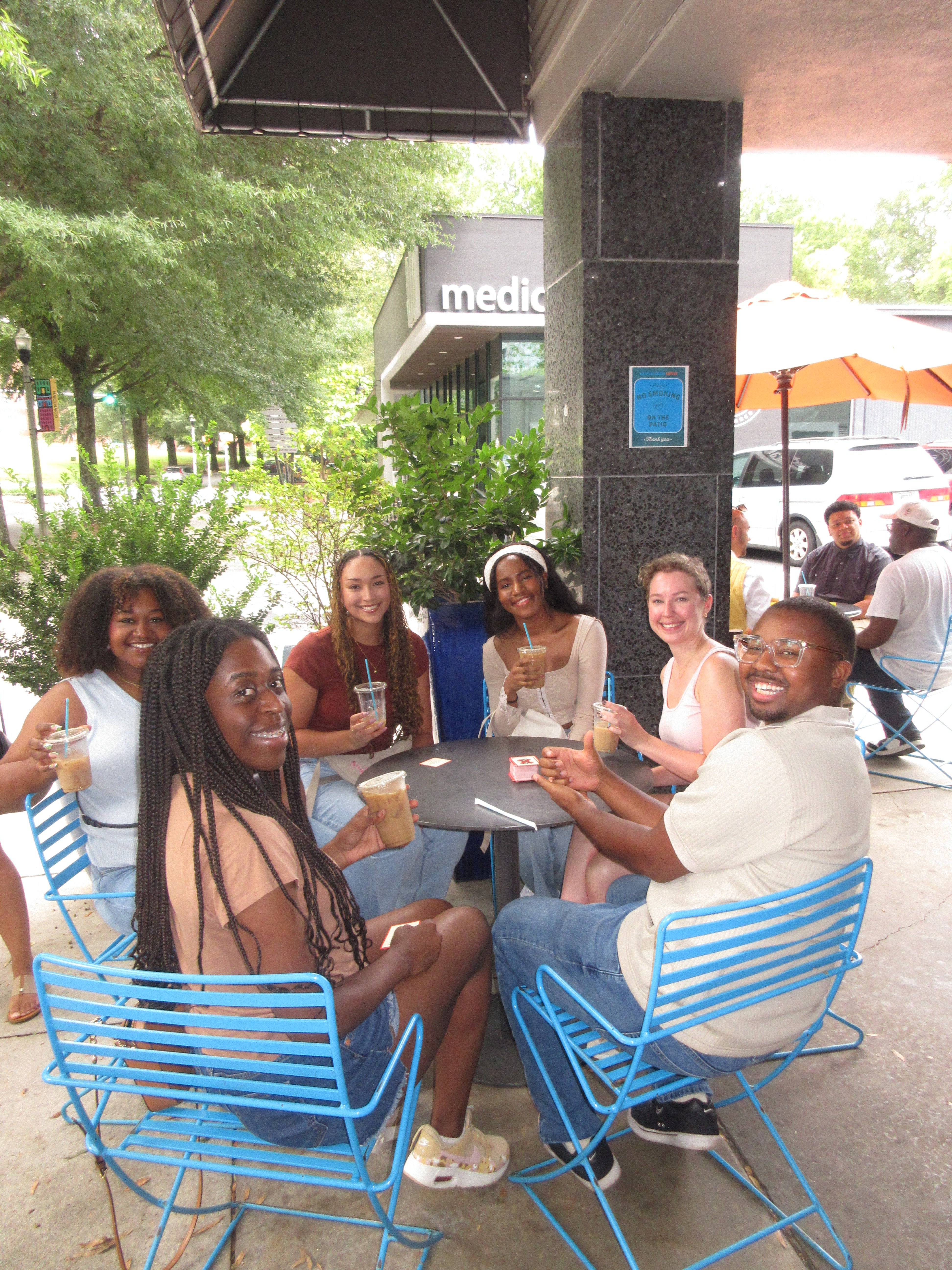 Group of people sitting at a table outdoors with blue chairs.