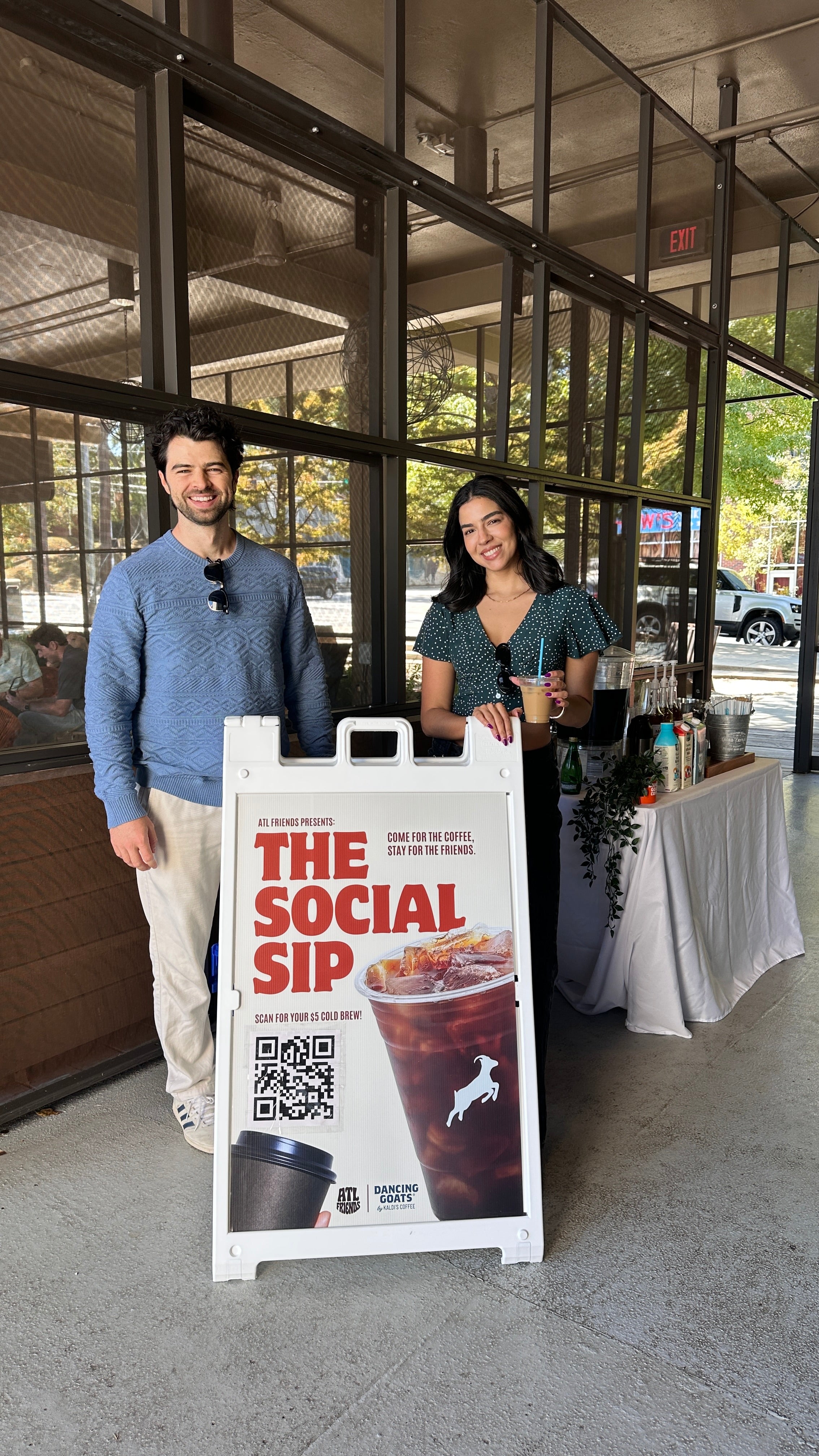 Two people standing next to a 'The Social Sip' sign in an outdoor setting.