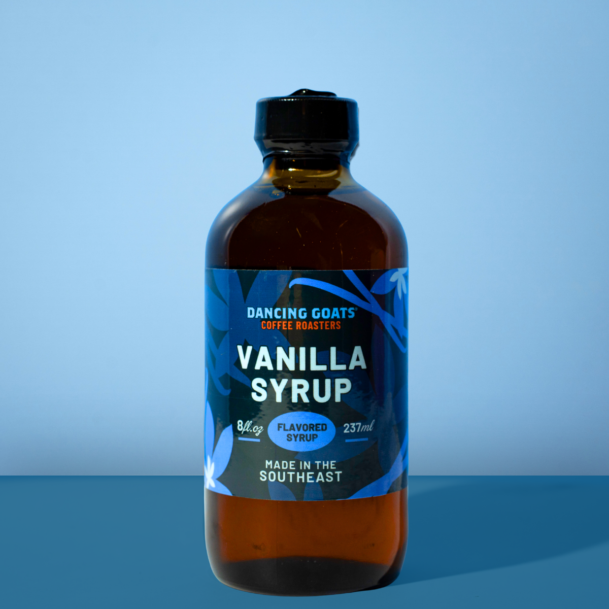 A bottle of Dancing Goats Coffee Roasters Vanilla Syrup in an amber glass bottle with a black cap and a blue label, set against a blue gradient background.