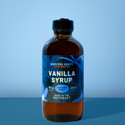 A bottle of Dancing Goats Coffee Roasters Vanilla Syrup in an amber glass bottle with a black cap and a blue label, set against a blue gradient background.