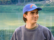 Person wearing a blue cap with a logo, standing by a body of water.