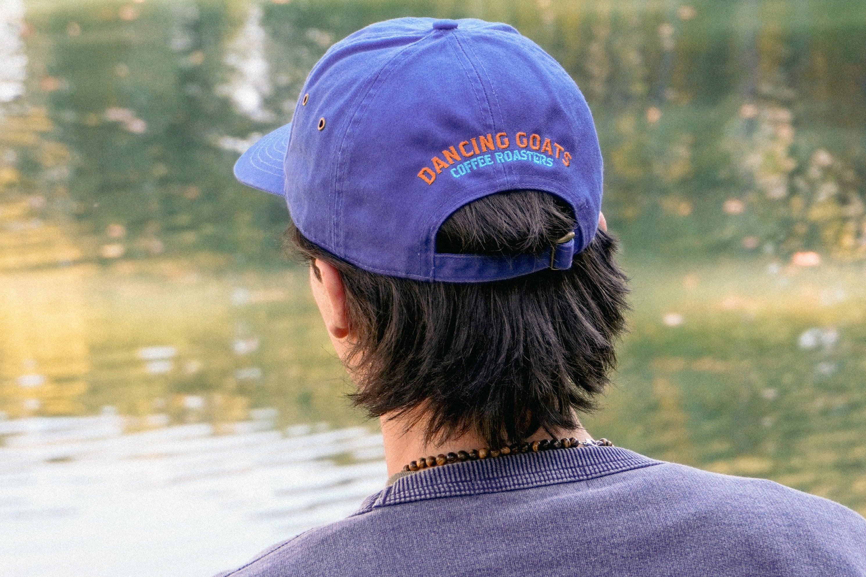 Person wearing a blue cap with 'Dancing Goats Coffee Roasters' logo, facing a body of water.